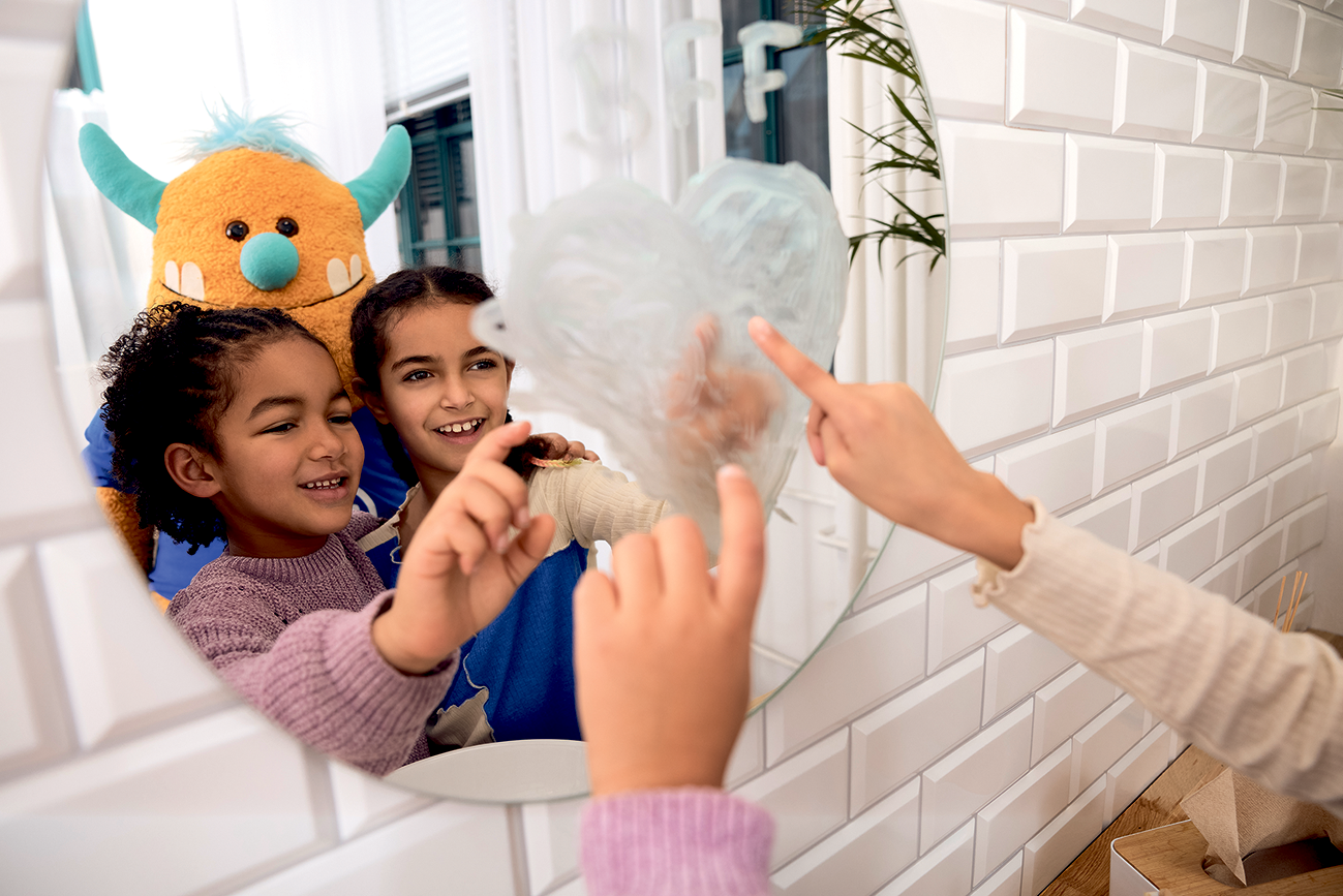 Zwei Kinder und Plüschmonster stehend vor Spiegel, auf den sie ein Herz mit Zahnpasta malen