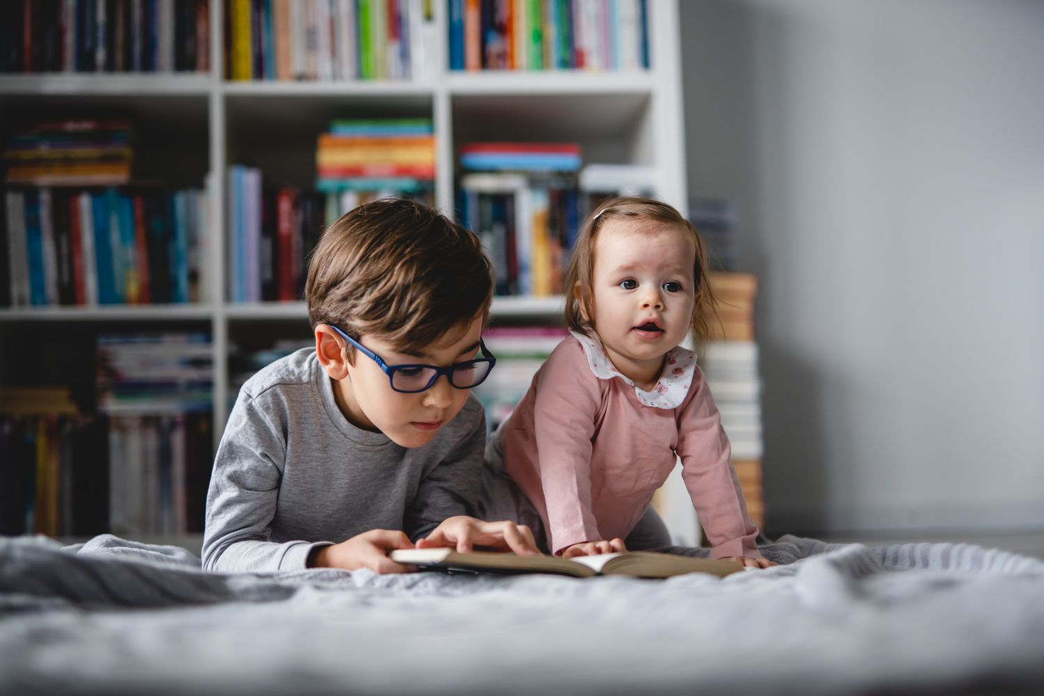 Zwei Kinder liegen auf einer Decke; ein Junge liest ein Buch, die Schwester schaut in die Gegend